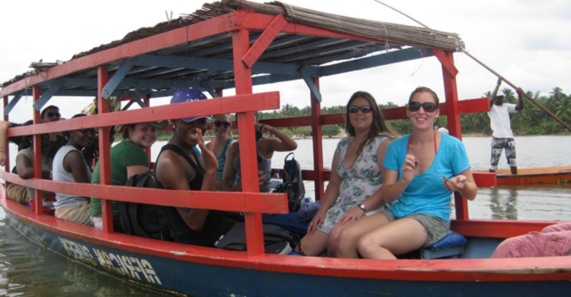 Students sit in a boat on the water and wave at the camera.